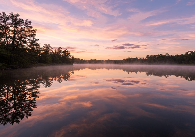 A photo of soft pastel clouds during sunrise reflected on a still lake with trees surrounding the water