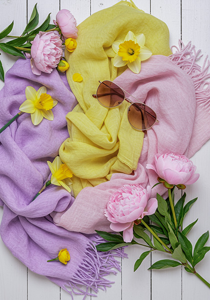 A photo of a flatlay of spring-themed accessories , soft pastel scarves, sunglasses, and flowers, arranged on a white wooden table