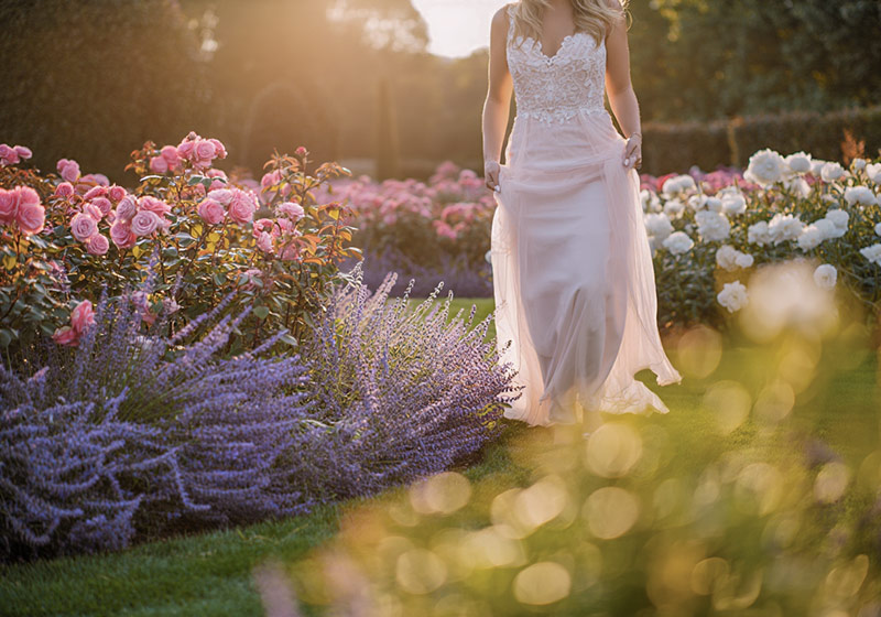 A photo of a bride standing in a sunlit garden surrounded by blooming pastel flowers, wearing a flowing white dress
