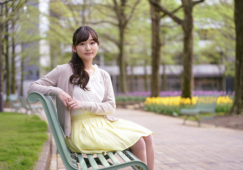 A photo of a young woman sitting on a pastel-colored bench in an urban park, wearing light spring clothes