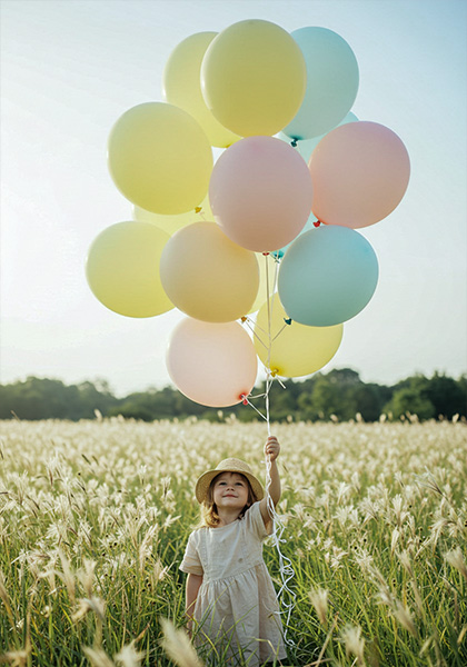 A child holding pastel-colored balloons in a field of light wildflowers. Balloons in pale lemon, blush pink, and sky blue fill the upper frame
