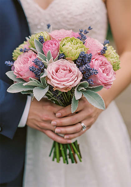 A photo of a wedding bouquet with muted pink, lavender, and sage-green flowers held by a bride