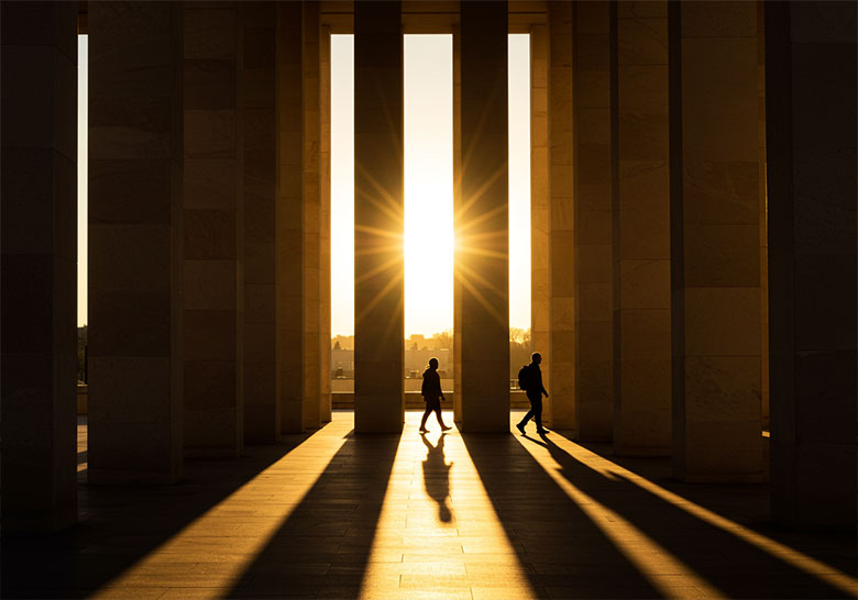 before - two persons walking between tall columns
